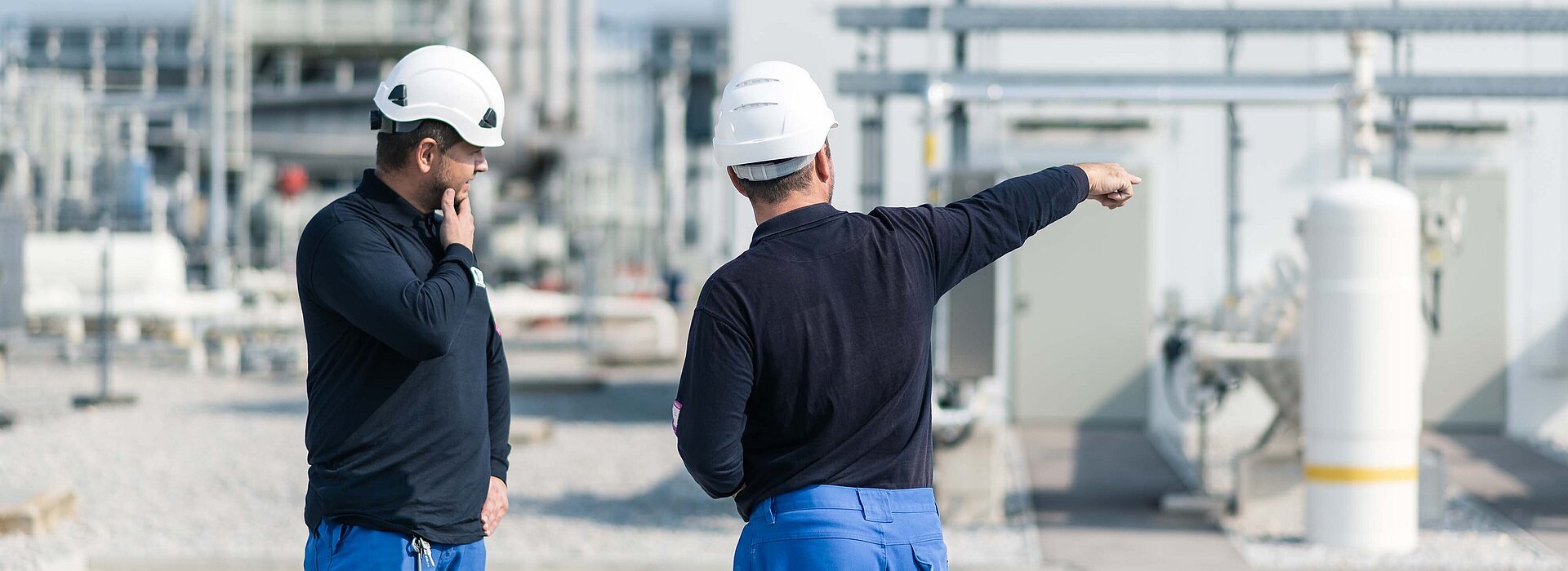 Zwei Montagemitarbeiter mit Sicherheitshelm, einer deutet auf etwas an der Industrieanlage im Hintergrund