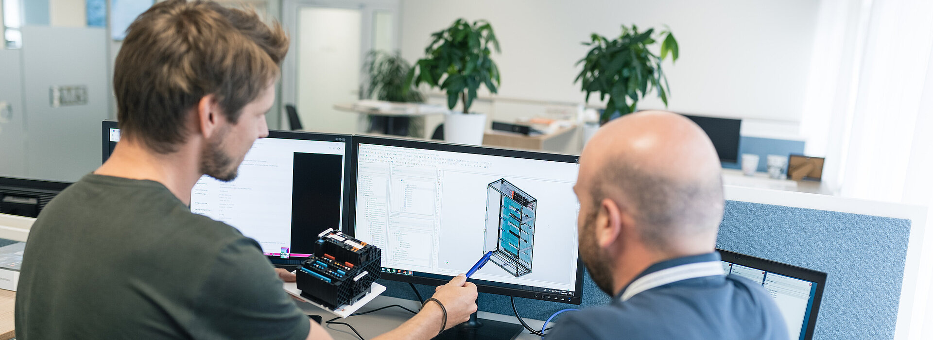 Zwei Automatisierungstechniker im Gespräch deuten auf eine Skizze am Bildschirm, dahinter das Großraumbüro