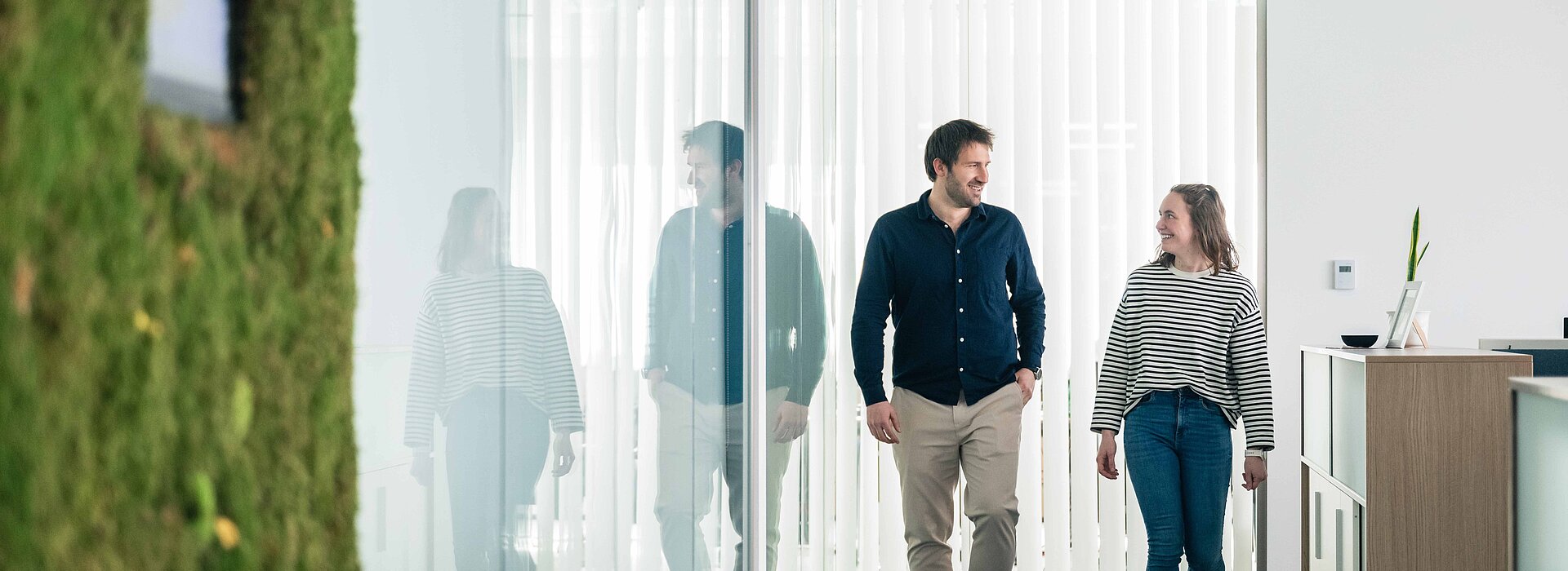 Ein Mann und eine Frau gehen einen Gang in einem Büro entlang und unterhalten sich, links sieht man eine grüne Mooswand