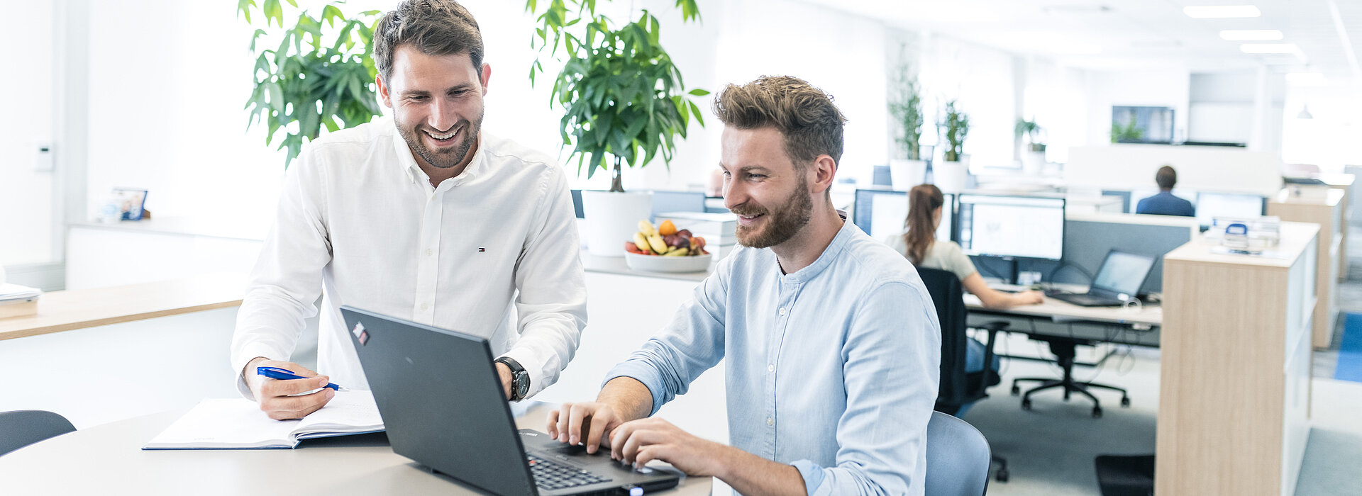 Zwei lächelnde Mitarbeiter der PSI beim Abstimmen mit Laptop und Block und Stift am Stehtisch, dahinter das moderne Großraumbüro mit arbeitenden Kolleg:innen