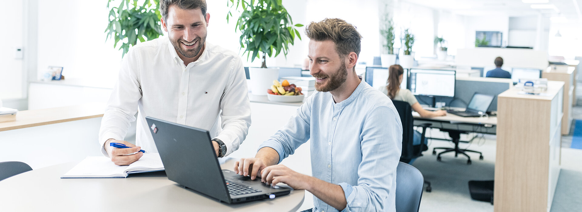 Zwei lächelnde Mitarbeiter der PSI beim Abstimmen mit Laptop und Block und Stift am Stehtisch, dahinter das moderne Großraumbüro mit arbeitenden Kolleg:innen