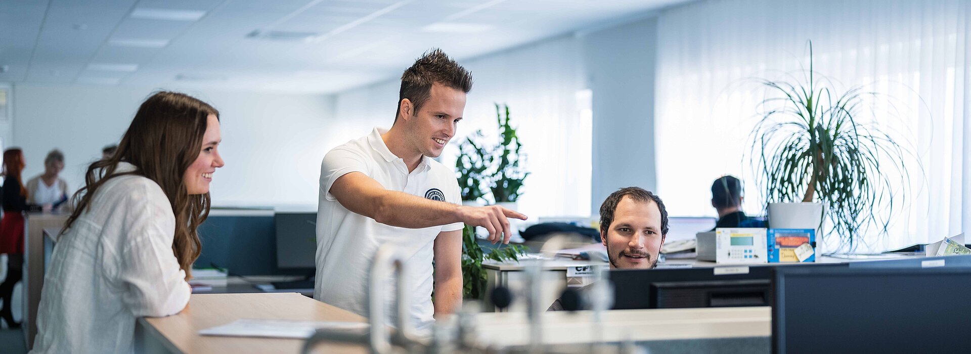 Mitarbeiter:innen im Großraumbüro diskutieren über einen PC gebeugt, davor ein technisches Teil am Tresen