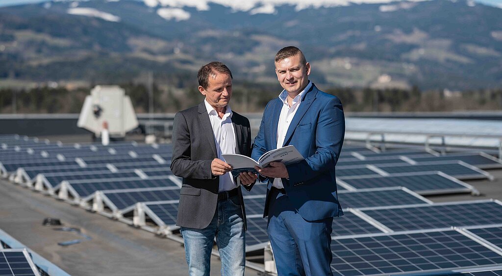 zwei Geschäftsführer am Hallendach, im Hintergrund die PV Anlage und schneebedeckte Berge