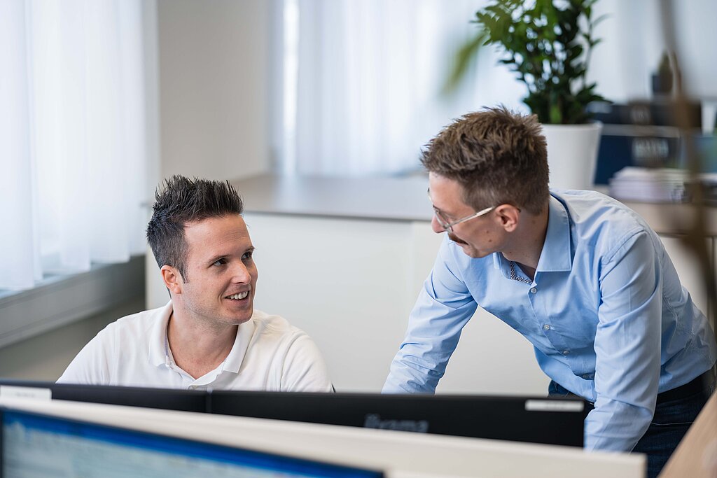 Ein am Schreibtisch sitzender Mitarbeiter bespricht etwas mit einem über den Schreibtisch gebeugten Kollegen