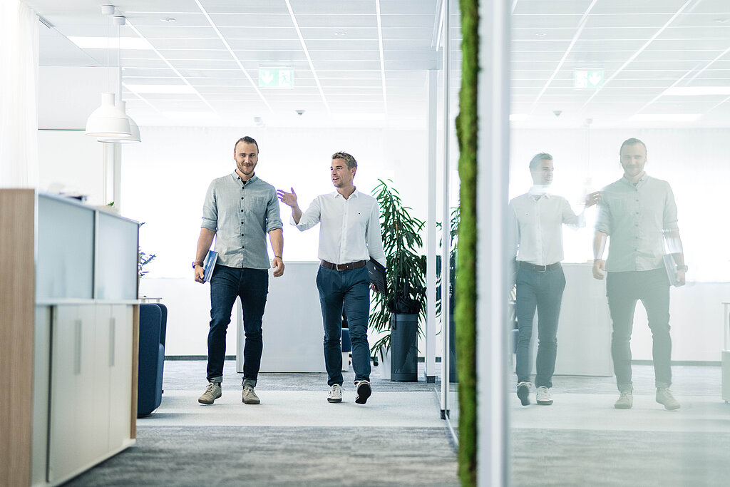 Zwei Mitarbeiter schreiten den Gang des Großraumbüros entlang und unterhalten sich