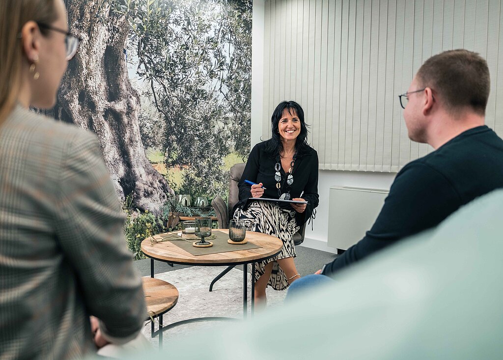Coachinggespräch mit zwei Mitarbeiten im PMS Coachingraum, auf der Tapete im Hintergrund ist ein Olivenbaum zu sehen