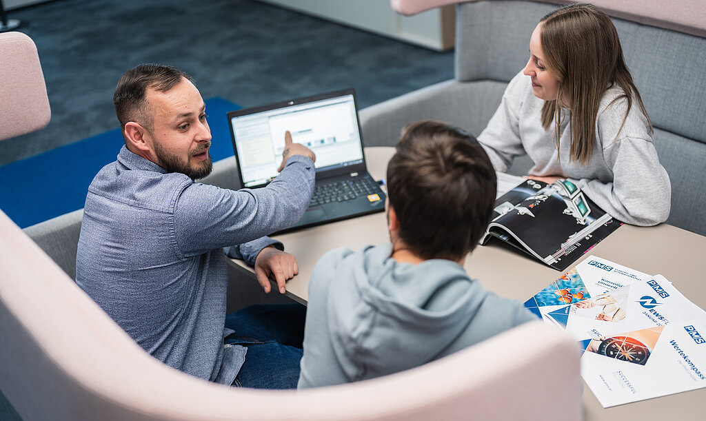 2 Mitarbeiter und ein Mitarbeiterin sitzen in der Sofaecke im Büro und besprechen sich mit Laptop und Unterlagen am Tisch