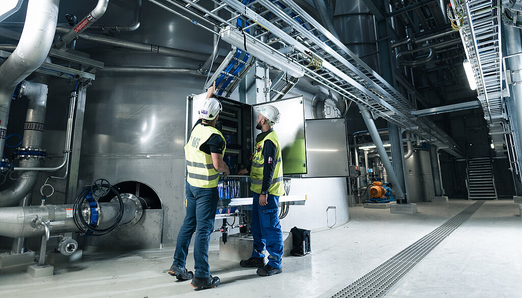 Zwei Montagemitarbeiter mit Sicherheitshelm und Warnweste stehen an einer Industrieanlage mit zahlreichen Kabeltrassen und einem geöffneten Schaltkasten