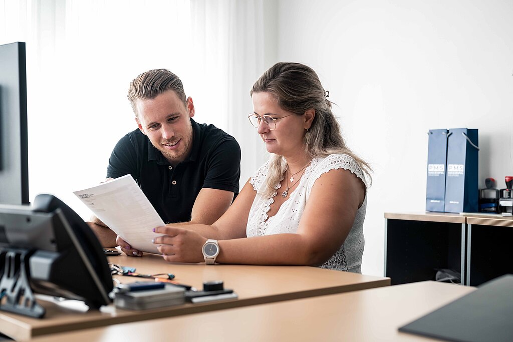 Zwei Mitarbeiter:innen beim Durchsehen einer Liste am Schreibtisch in einem modernen Büro
