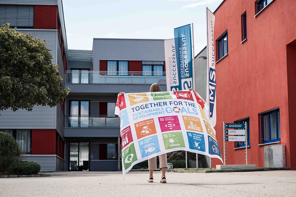 Eine Mitarbeiterin mit einer SDG Fahne vor dem Eingang des Firmengebäudes der PMS