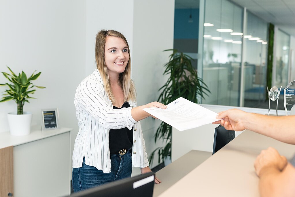 Eine Mitarbeiterin am Empfang übergibt Unterlagen an eine weitere Person vor dem Empfangstisch