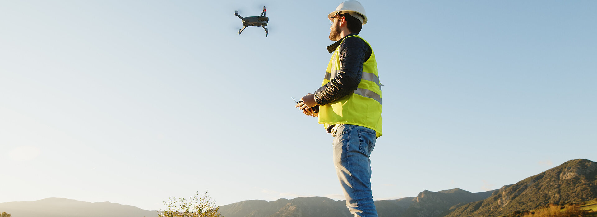Ein Mann mit Helm und Sicherheitswese steuert eine Drohne auf einer Alm in den Bergen