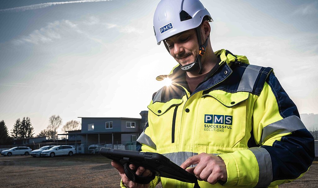 Ein Arbeiter in Sicherheitsjacke und Helm am Tablett, dahinter das Bürogebäude mit der untergehenden Sonne