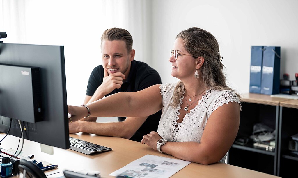 Mitarbeiter der PSI deuten auf einen Bildschirm, beide stehend am höhenverstellbaren Schreibtisch
