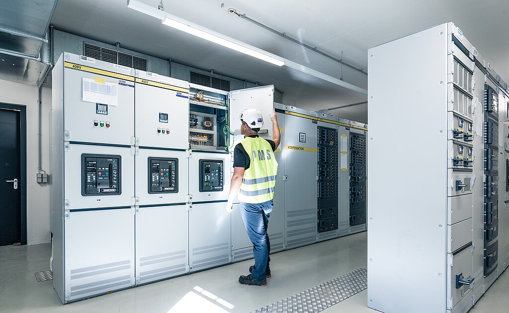 PMS Mitarbeiter mit Sicherheitshelm und Warnweste öffnet eine Tür in einer Reihe von Schaltschränken