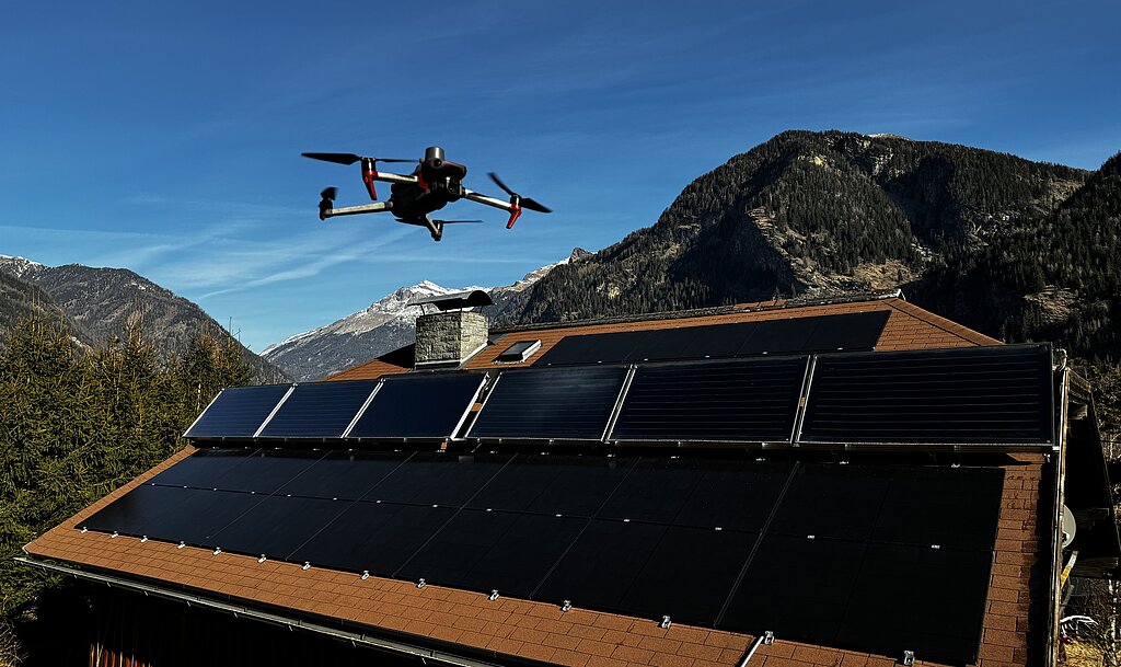 Eine Drohne fliegt über ein Gebäudedach mit PV Anlage, im Hintergrund schneebedeckte Berge