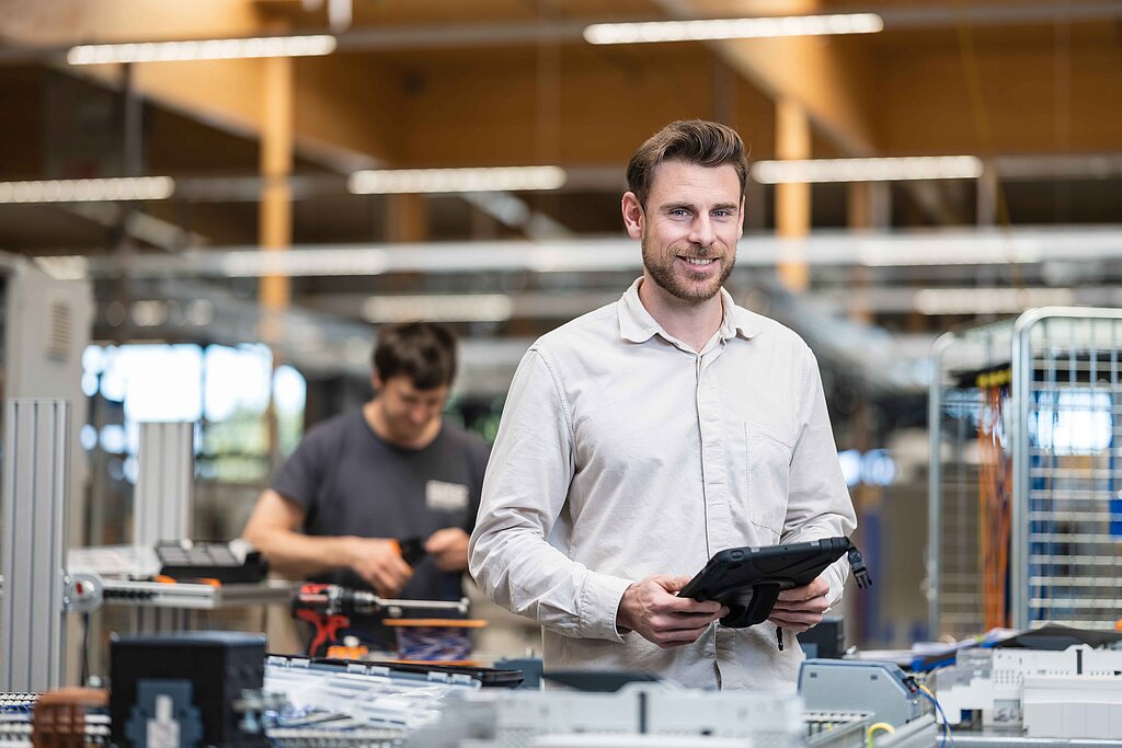 CCS Führungskraft mit Tablet in der Fertigungshalle, im Hintergrund zahlreiche Schaltschränke und arbeitende Kollegen